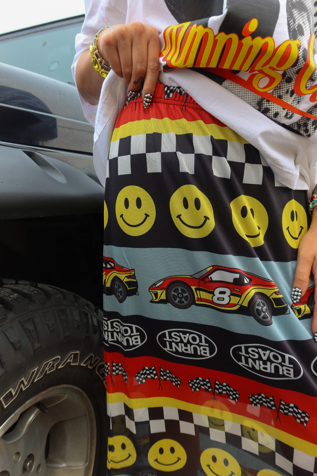 A close-up of a person wearing patterned athletic shorts next to a car tire. The shorts feature yellow smiley faces, checkered stripes, red and yellow race cars, and 'Burnt Toast' text