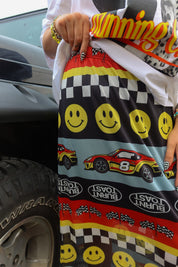 A close-up of a person wearing patterned athletic shorts next to a car tire. The shorts feature yellow smiley faces, checkered stripes, red and yellow race cars, and 'Burnt Toast' text