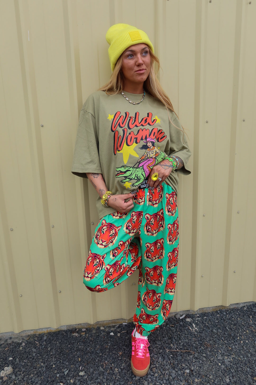 A woman stands against a beige corrugated metal wall wearing a bright yellow beanie, an oversized olive green graphic T-shirt that reads 'Wild Woman', green pants covered in a red tiger print, and pink sneakers