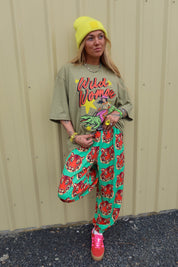 A woman stands against a beige corrugated metal wall wearing a bright yellow beanie, an oversized olive green graphic T-shirt that reads 'Wild Woman', green pants covered in a red tiger print, and pink sneakers