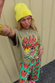 A woman takes a selfie wearing a bright yellow knit beanie, a distressed olive green graphic t-shirt with 'Wild Woman' text, and green pants with a red tiger print, standing against a beige corrugated metal wall