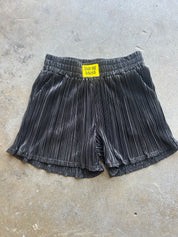 Black pleated shorts with a yellow 'burnt toast!' label at the waistband, laid flat on a gray concrete floor
