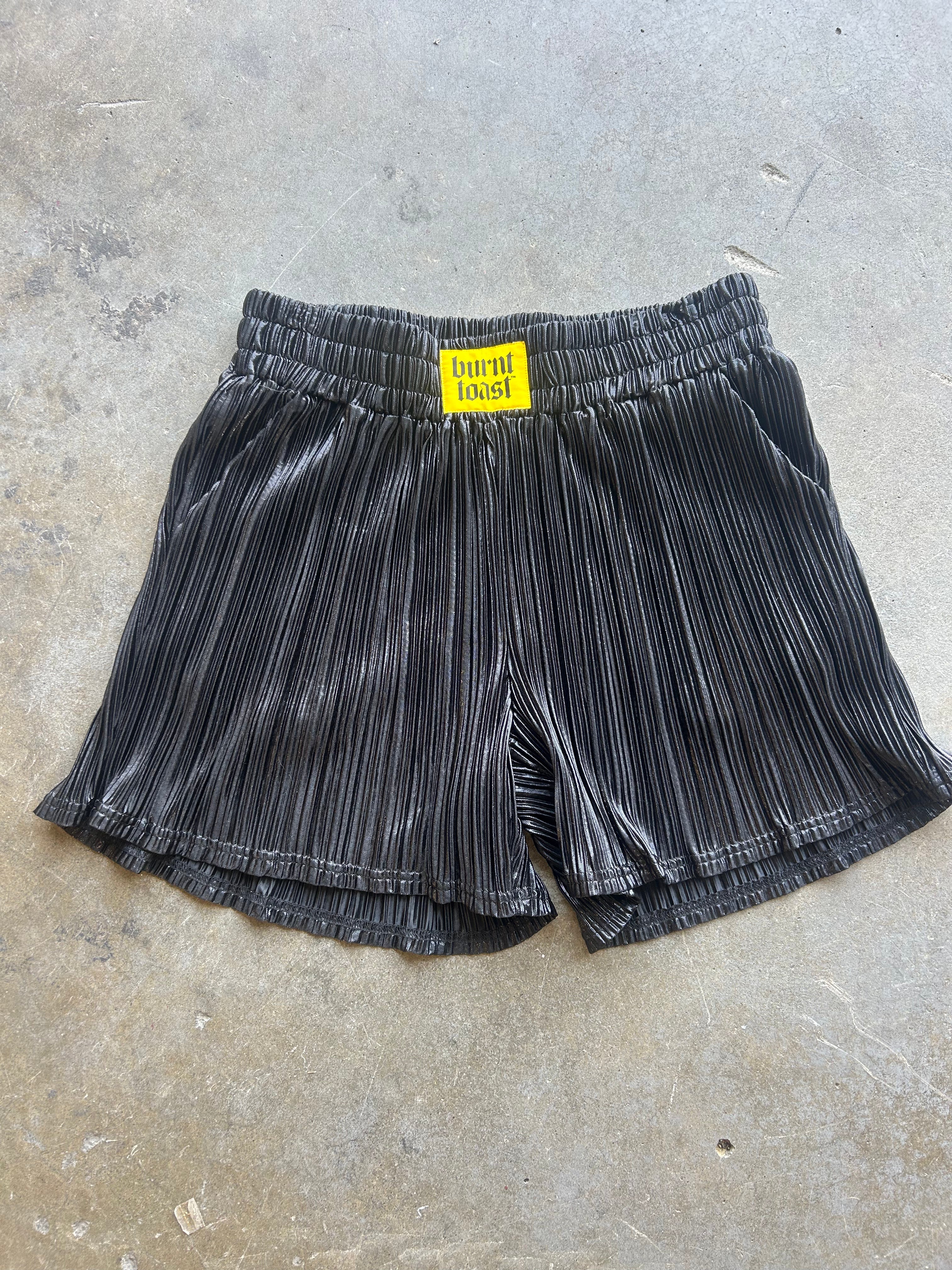 Black pleated shorts with a yellow 'burnt toast!' label at the waistband, laid flat on a gray concrete floor