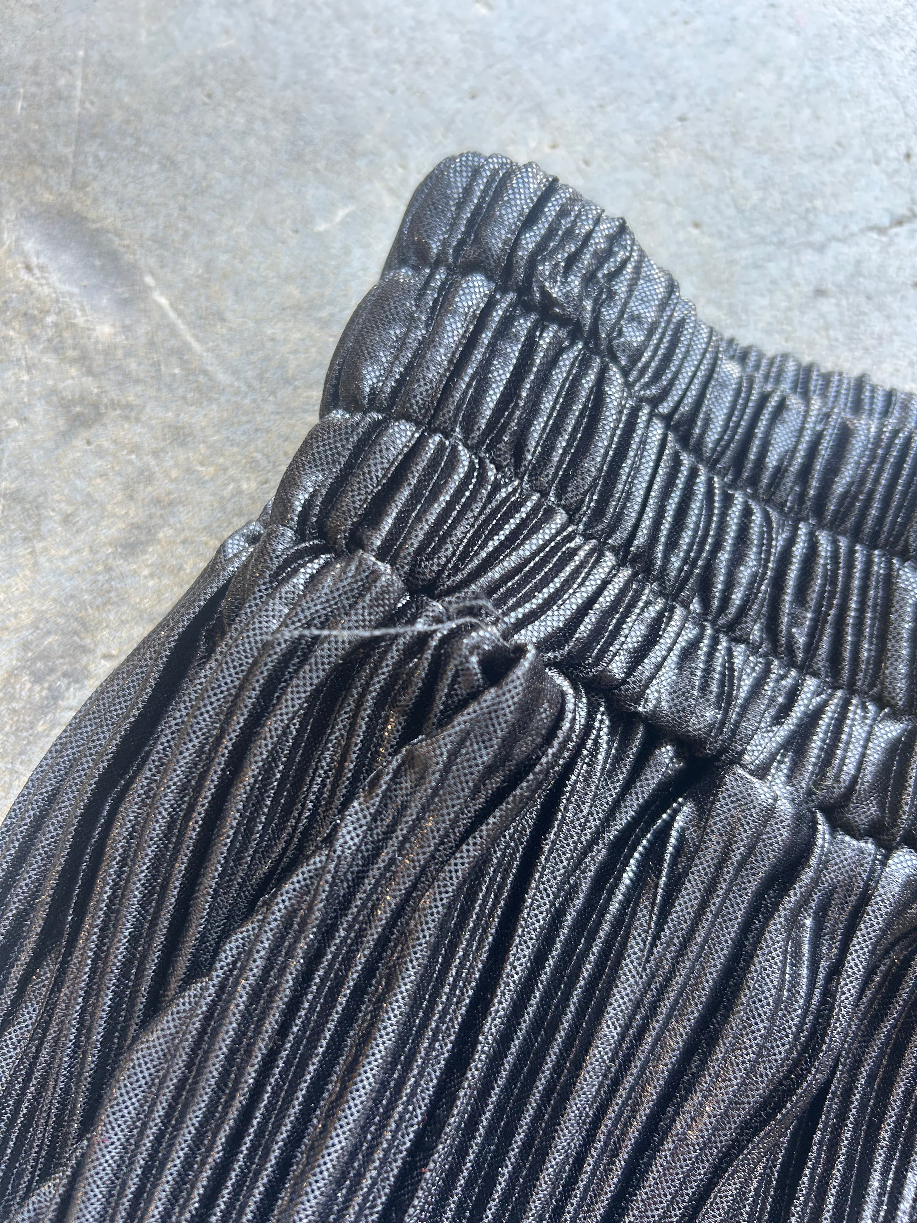 Close-up of a black pleated skirt with a gathered elastic waistband, laid flat on a gray concrete surface