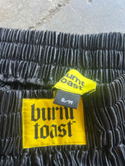 Close-up of black textured shorts with a yellow Burnt Toast brand label and a small black size tag reading S/M