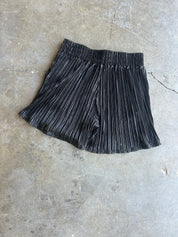 Black pleated shorts with a gathered elastic waistband, laid flat on a textured gray concrete surface