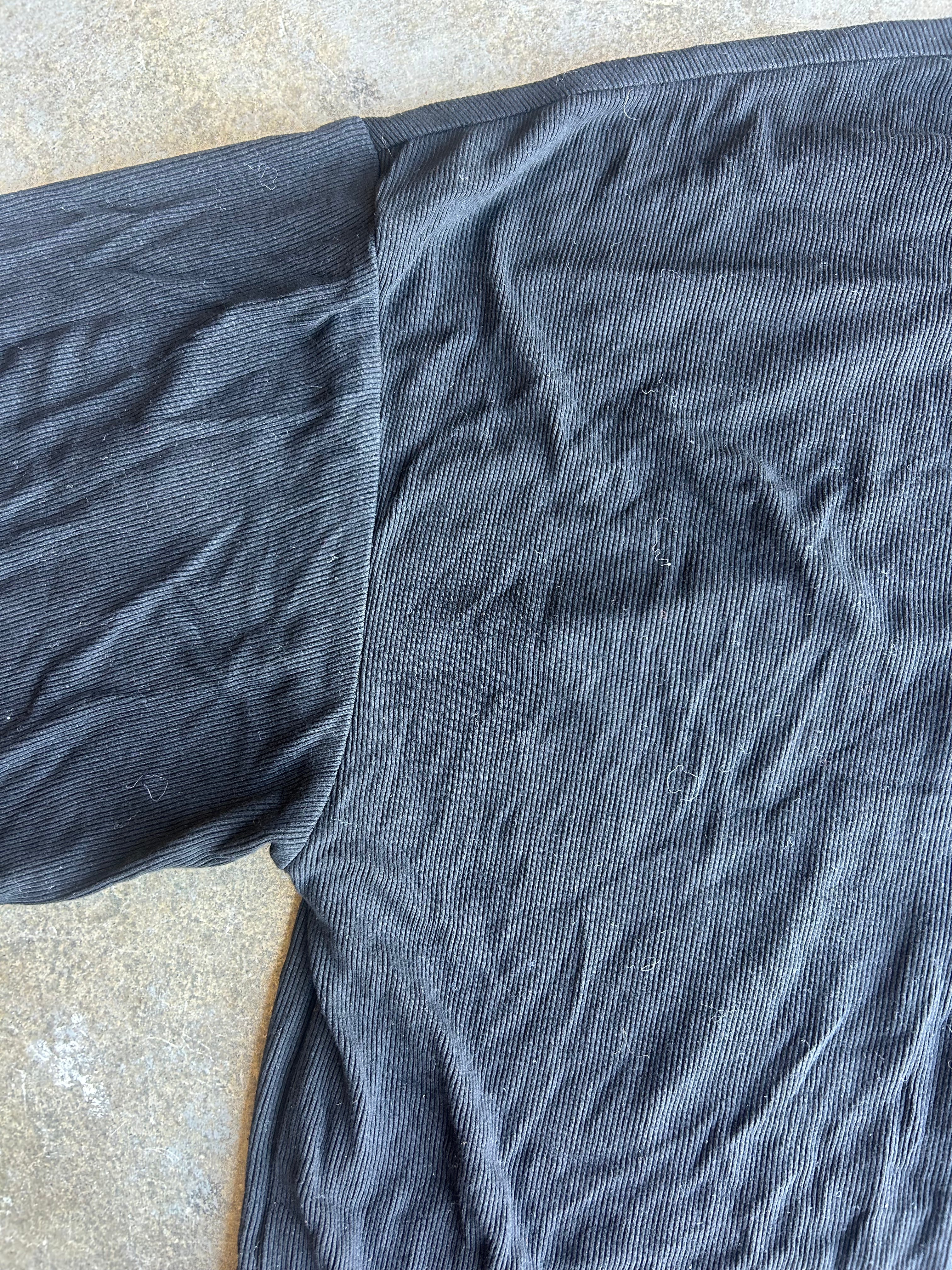 A close-up of a dark charcoal gray ribbed knit fabric, likely a t-shirt body, laid flat on a textured concrete surface