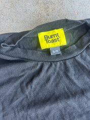 Close-up of a dark gray ribbed t-shirt with a bright yellow Burnt Toast brand neck label and a small gray size tag reading S/M, laid flat on a textured concrete surface