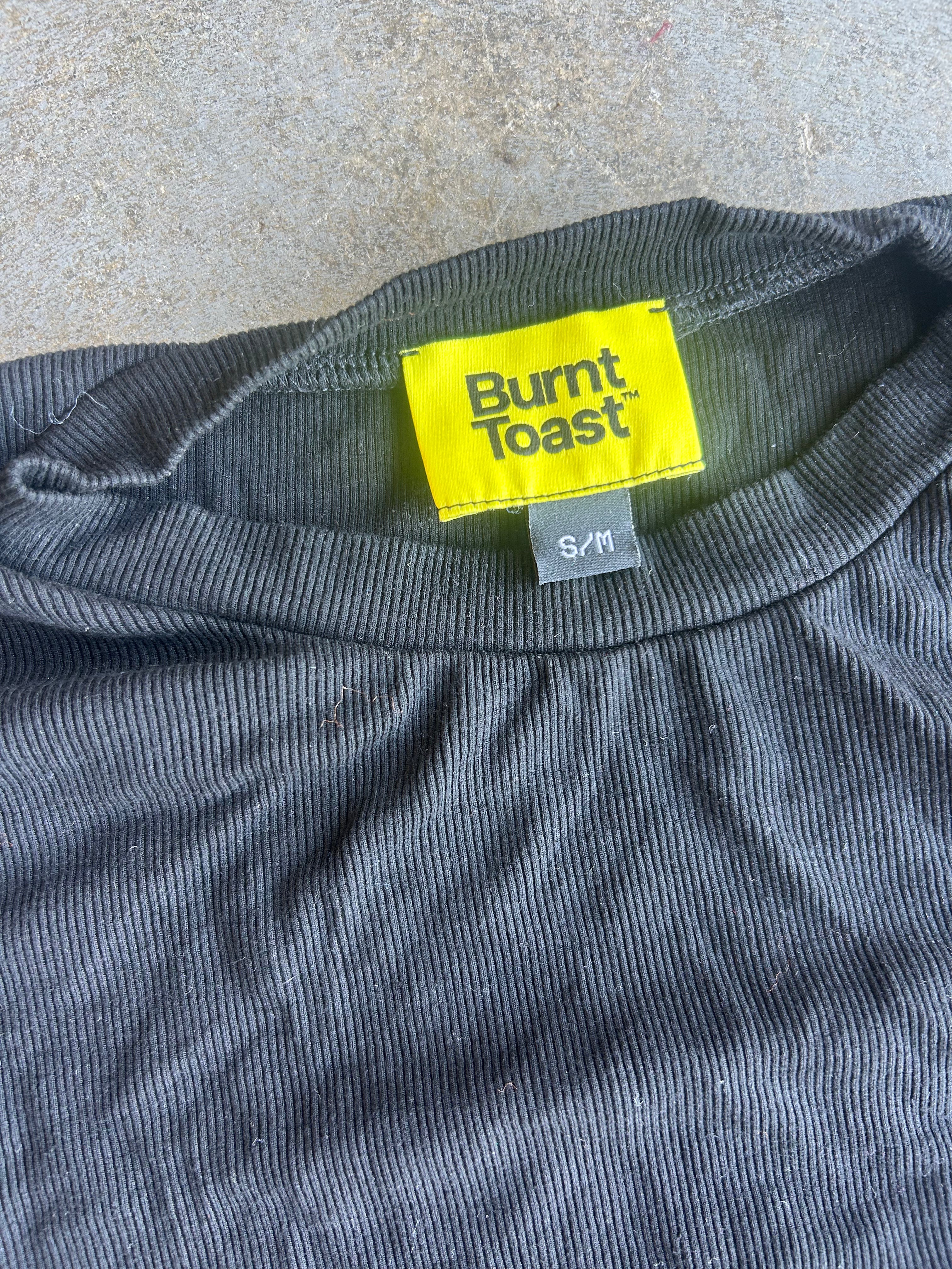 Close-up of a dark gray ribbed t-shirt with a bright yellow Burnt Toast brand neck label and a small gray size tag reading S/M, laid flat on a textured concrete surface