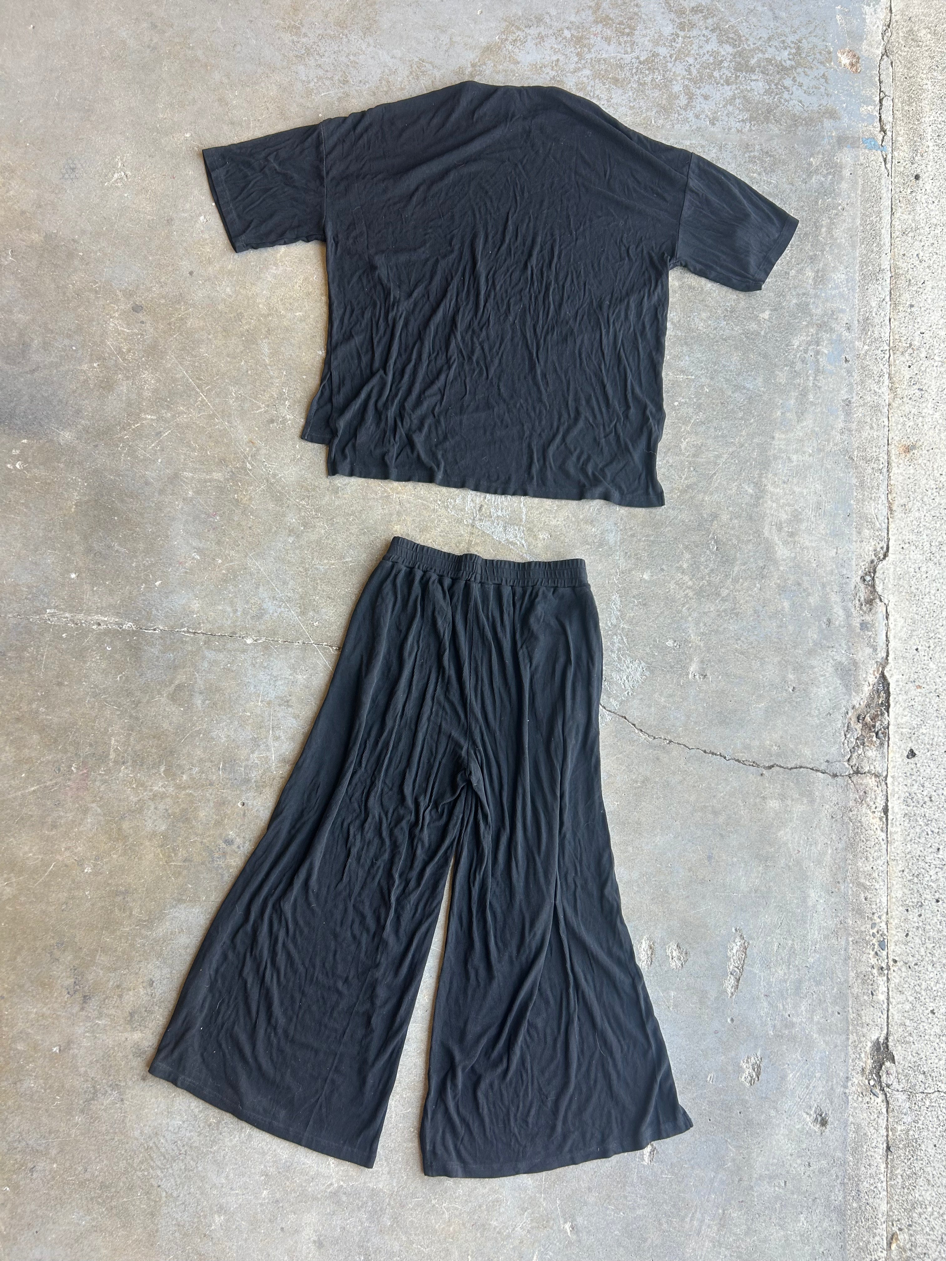 A black short-sleeve cropped T-shirt and matching black wide-leg pants laid flat on a textured concrete floor