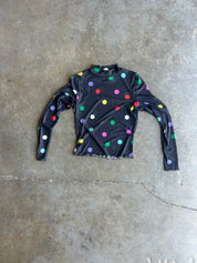 A black long-sleeve mock neck shirt with colorful polka dots, laid flat on a gray concrete floor