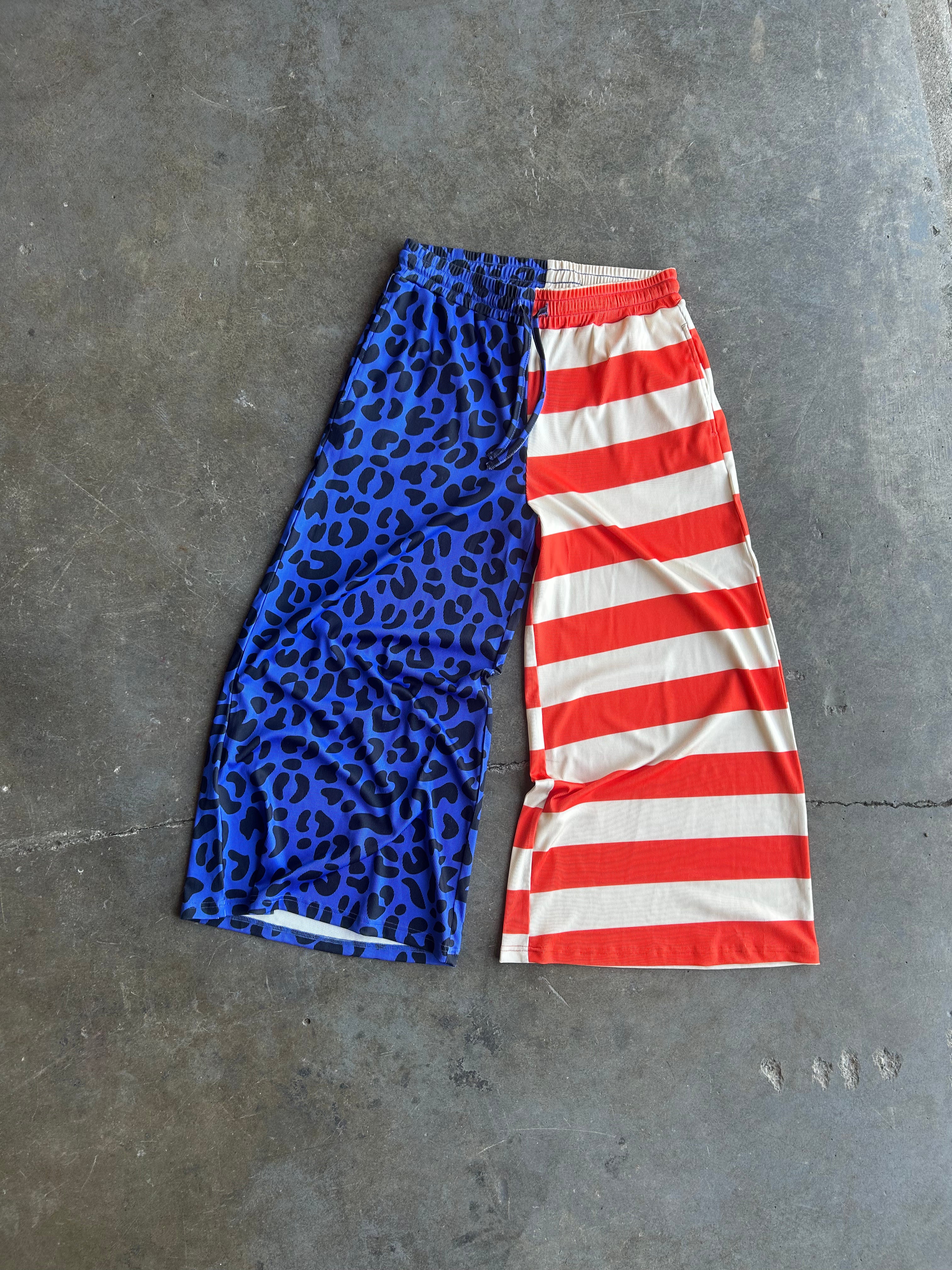 A pair of cropped athletic shorts laid flat on a gray concrete floor, featuring a split design: one half has a blue leopard print pattern and the other half has red and white horizontal stripes
