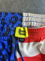 Close-up of a pair of shorts with a red, white, and blue patriotic design. The left side features a blue and black abstract pattern, the center has a blue-stitched cream and red striped design, and the right side is solid red