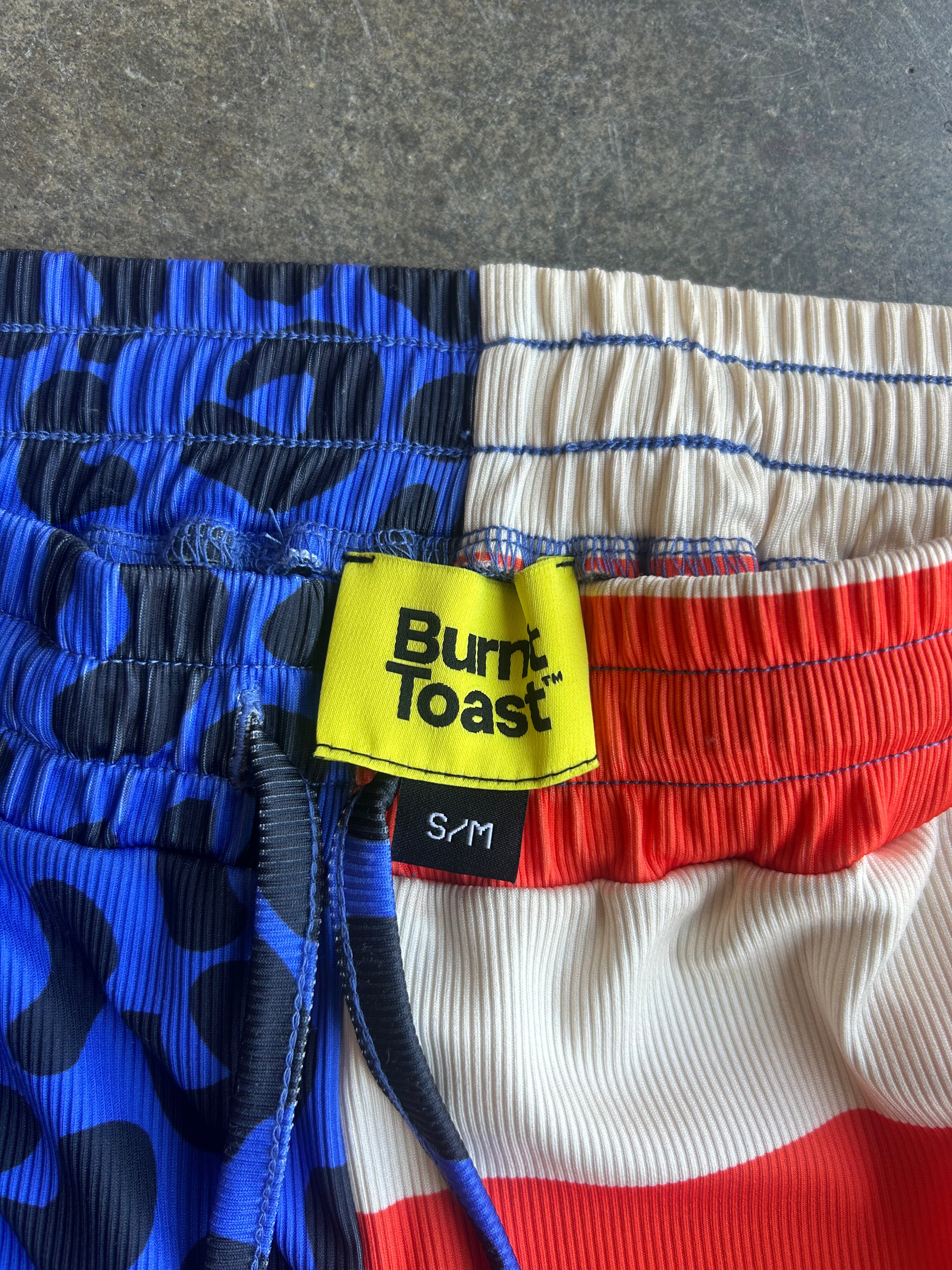 Close-up of a pair of shorts with a red, white, and blue patriotic design. The left side features a blue and black abstract pattern, the center has a blue-stitched cream and red striped design, and the right side is solid red
