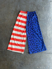 A pair of wide-leg cropped pants laid flat on a gray concrete surface, featuring a split design: one leg has red and white horizontal stripes, and the other leg has a blue leopard print pattern with black spots