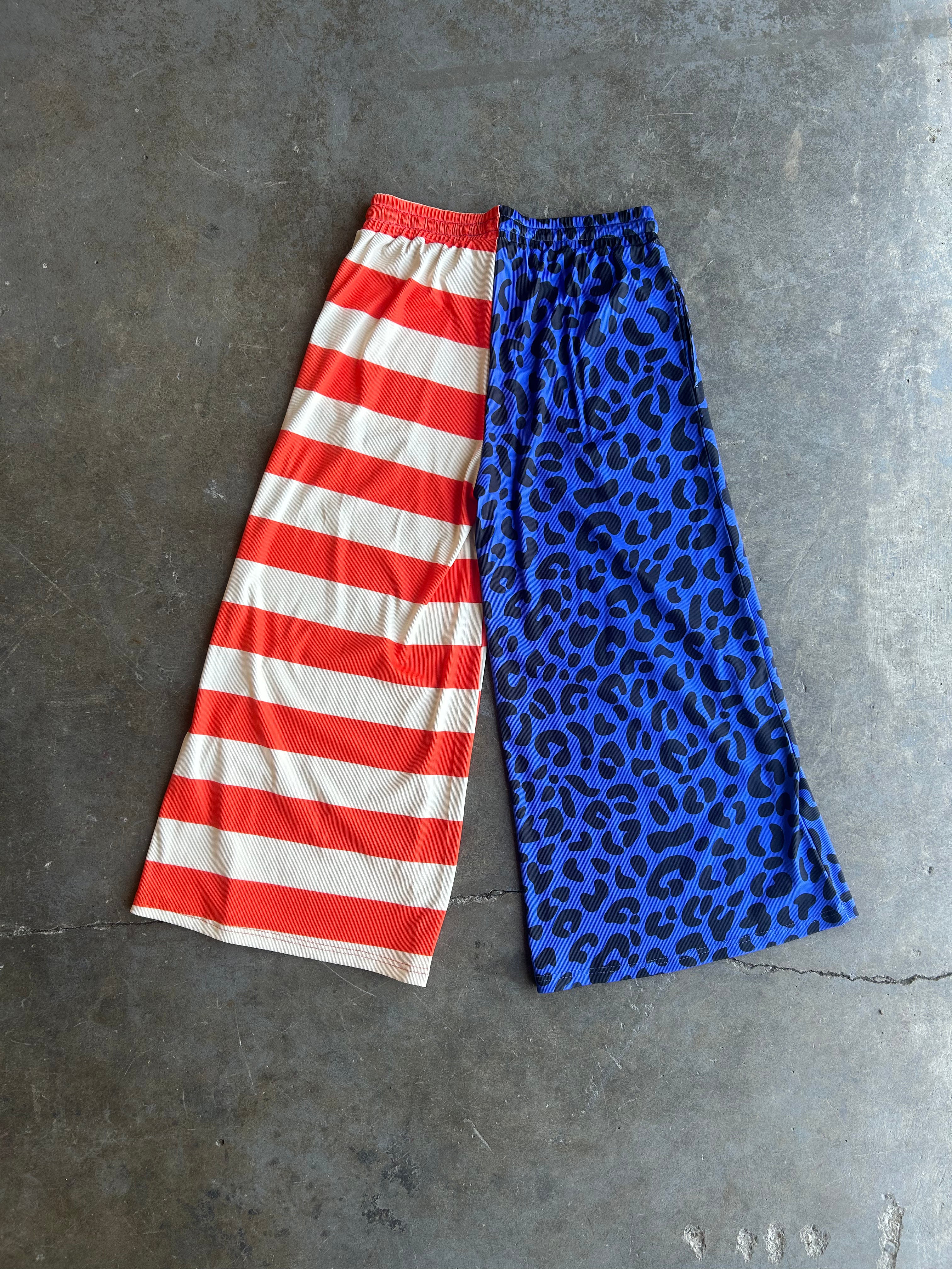 A pair of wide-leg cropped pants laid flat on a gray concrete surface, featuring a split design: one leg has red and white horizontal stripes, and the other leg has a blue leopard print pattern with black spots