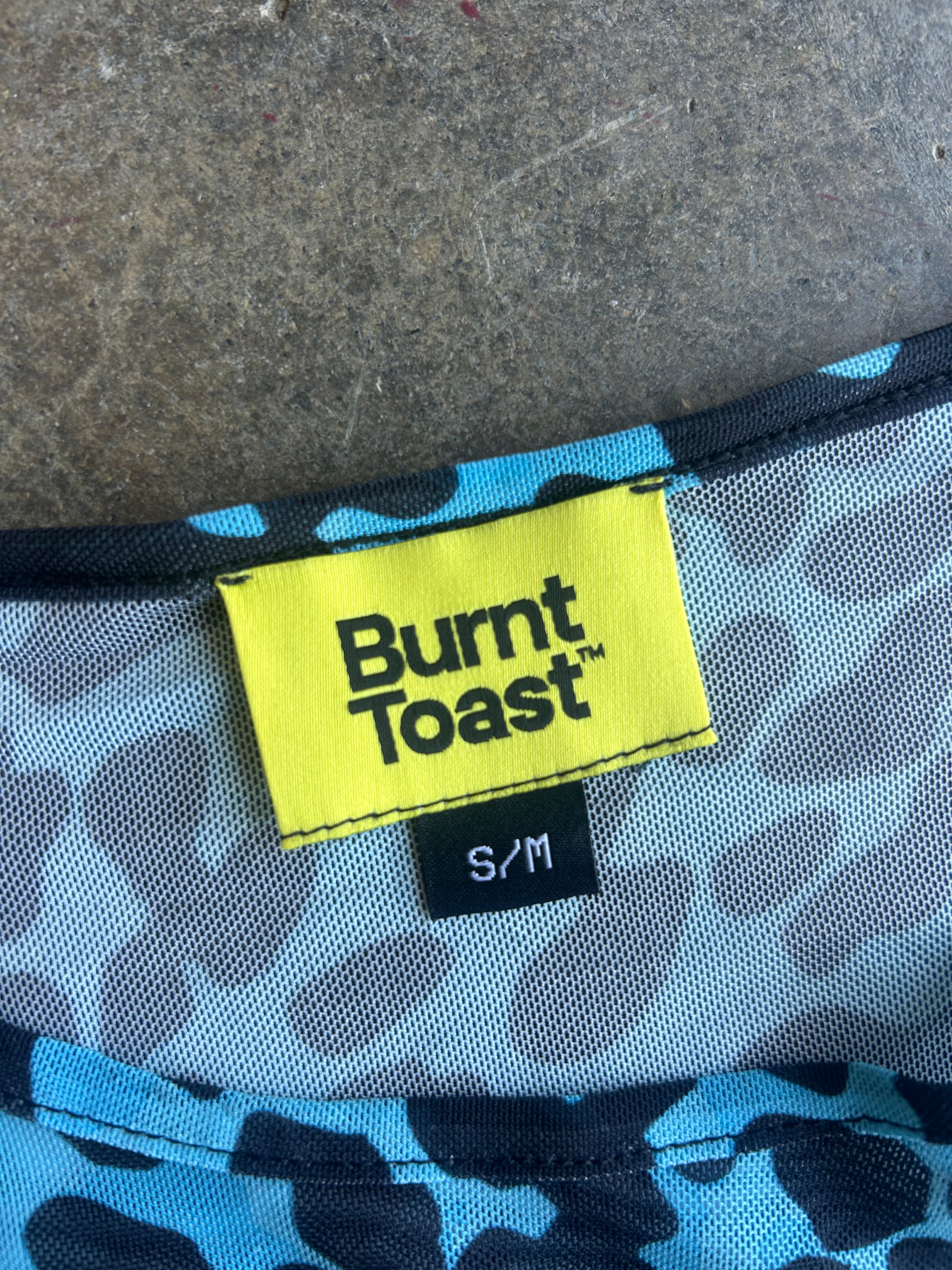 Close-up of a garment's inner neck label showing the brand 'Burnt Toast' in black text on a bright yellow tag, with a blue and gray patterned lining and a black size tag reading 'S/M'