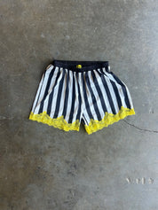 Black and white vertically striped shorts with a bright yellow lace hem and waistband detail, laid flat on a gray concrete floor