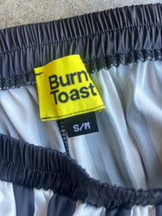 Close-up of a garment's waistband showing a bright yellow 'Burn Toast' brand label and a black 'S/M' size tag, with dark gray and white striped elastic trim