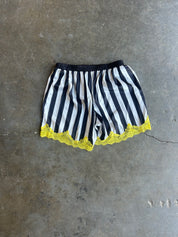 Black and white vertically striped shorts with a black elastic waistband and a bright yellow lace hem, laid flat on a gray concrete floor
