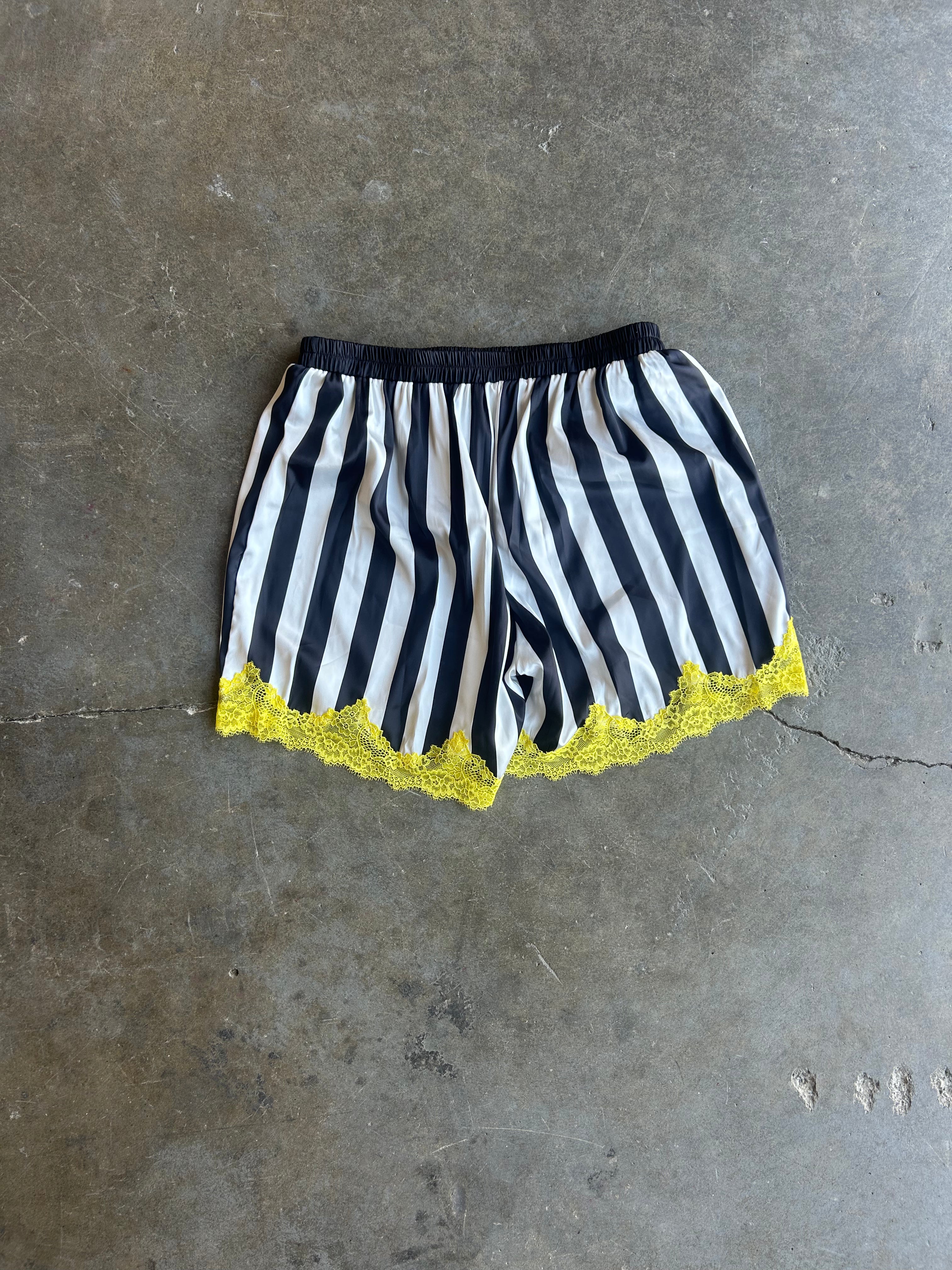 Black and white vertically striped shorts with a black elastic waistband and a bright yellow lace hem, laid flat on a gray concrete floor