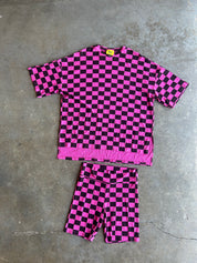 A two-piece matching lounge set consisting of a short-sleeve crewneck top and shorts, both featuring a vibrant pink and black checkerboard pattern, laid flat on a gray concrete floor