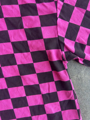 A close-up of a pink and black checkered garment, likely a sweatshirt or jacket, made from ribbed corduroy-like fabric, laid flat on a gray surface