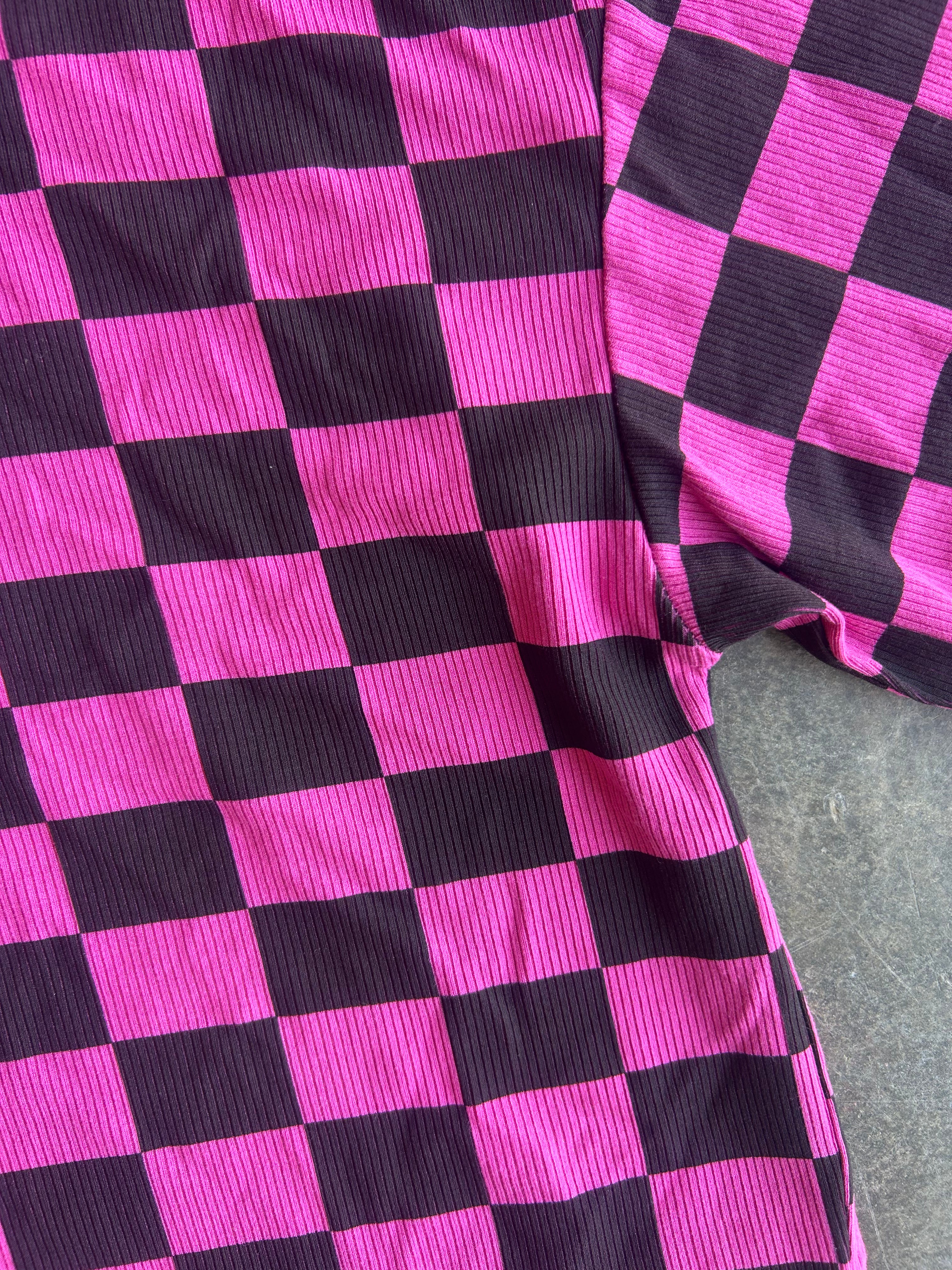 A close-up of a pink and black checkered garment, likely a sweatshirt or jacket, made from ribbed corduroy-like fabric, laid flat on a gray surface