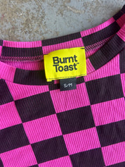 Close-up of a ribbed knit top with a hot pink and dark purple checkerboard pattern, featuring a bright yellow Burnt Toast brand label