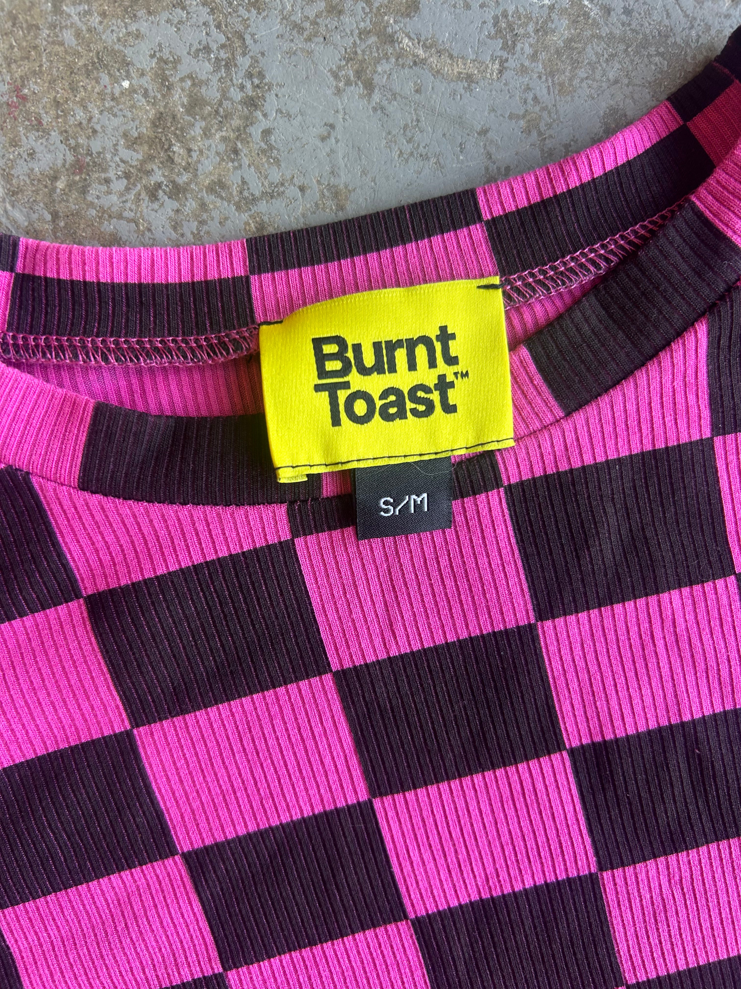 Close-up of a ribbed knit top with a hot pink and dark purple checkerboard pattern, featuring a bright yellow Burnt Toast brand label