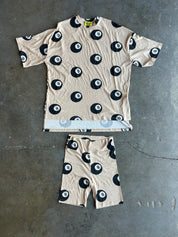 A matching short-sleeve t-shirt and shorts set laid flat on a gray concrete floor. The set features a beige base with a black and white 8-ball pattern, with a light blue and white striped hem detail on the shirt