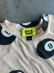 Close-up of a ribbed knit t-shirt with a black and white 8-ball pattern on a beige background, featuring a bright yellow Burnt Toast brand label