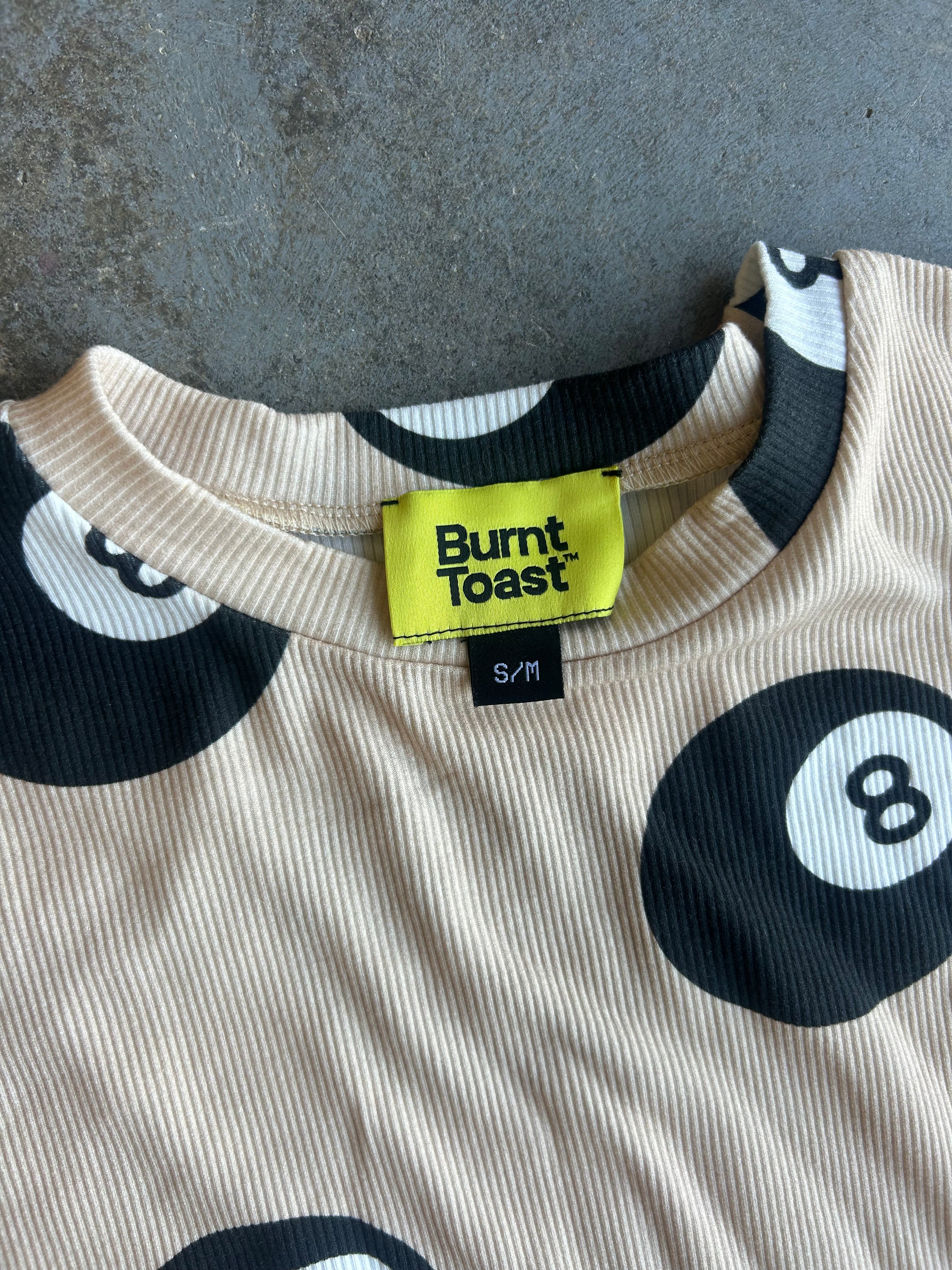 Close-up of a ribbed knit t-shirt with a black and white 8-ball pattern on a beige background, featuring a bright yellow Burnt Toast brand label