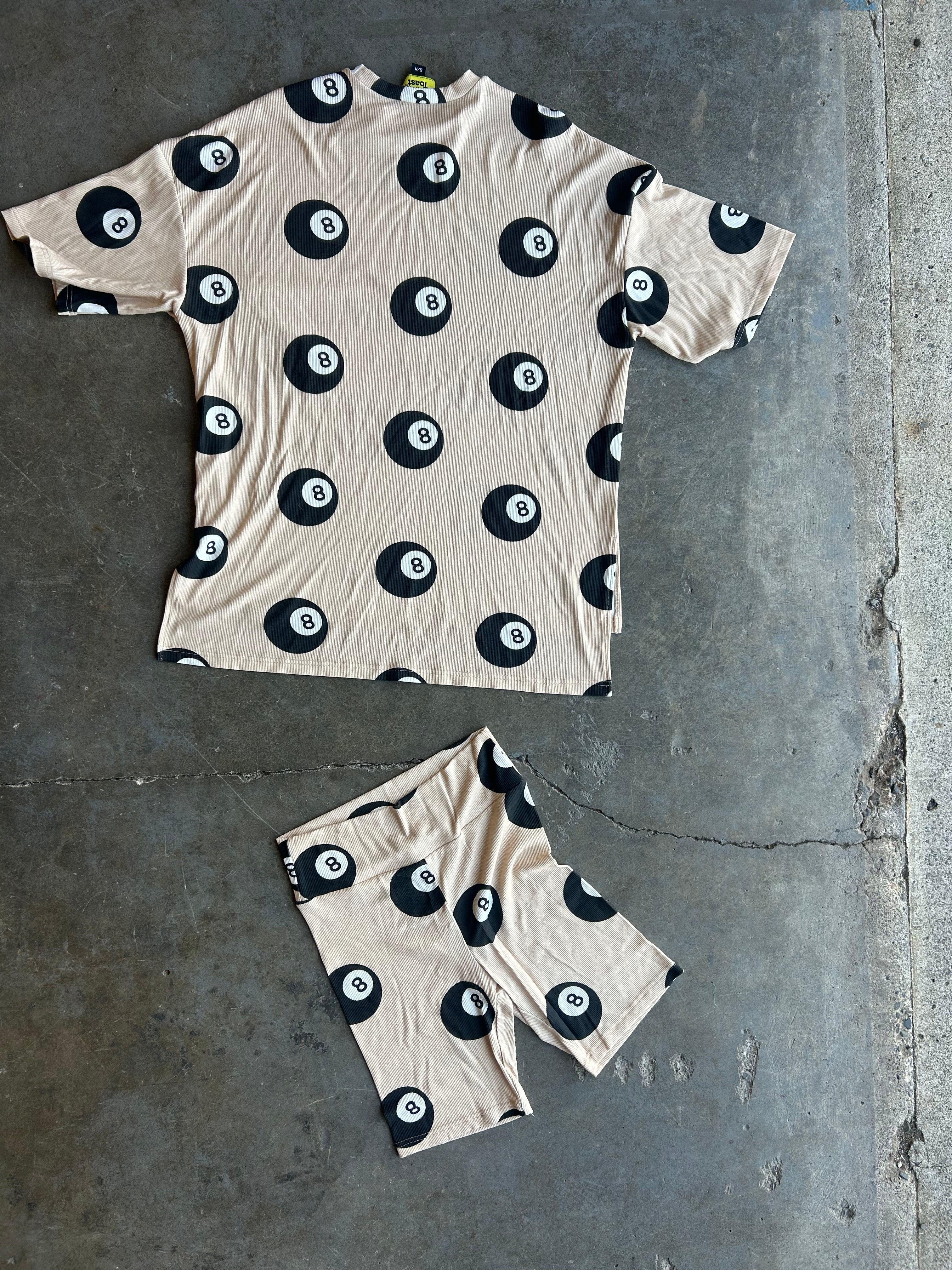 A matching beige short-sleeve t-shirt and shorts set with a black 8-ball pool pattern, laid flat on a gray concrete floor