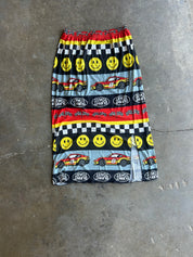 A knee-length skirt with an elastic waistband, featuring a colorful racing-themed print with yellow smiley faces, red race cars, checkered flags, and 'I'll Never Love' text, laid flat on a gray concrete floor