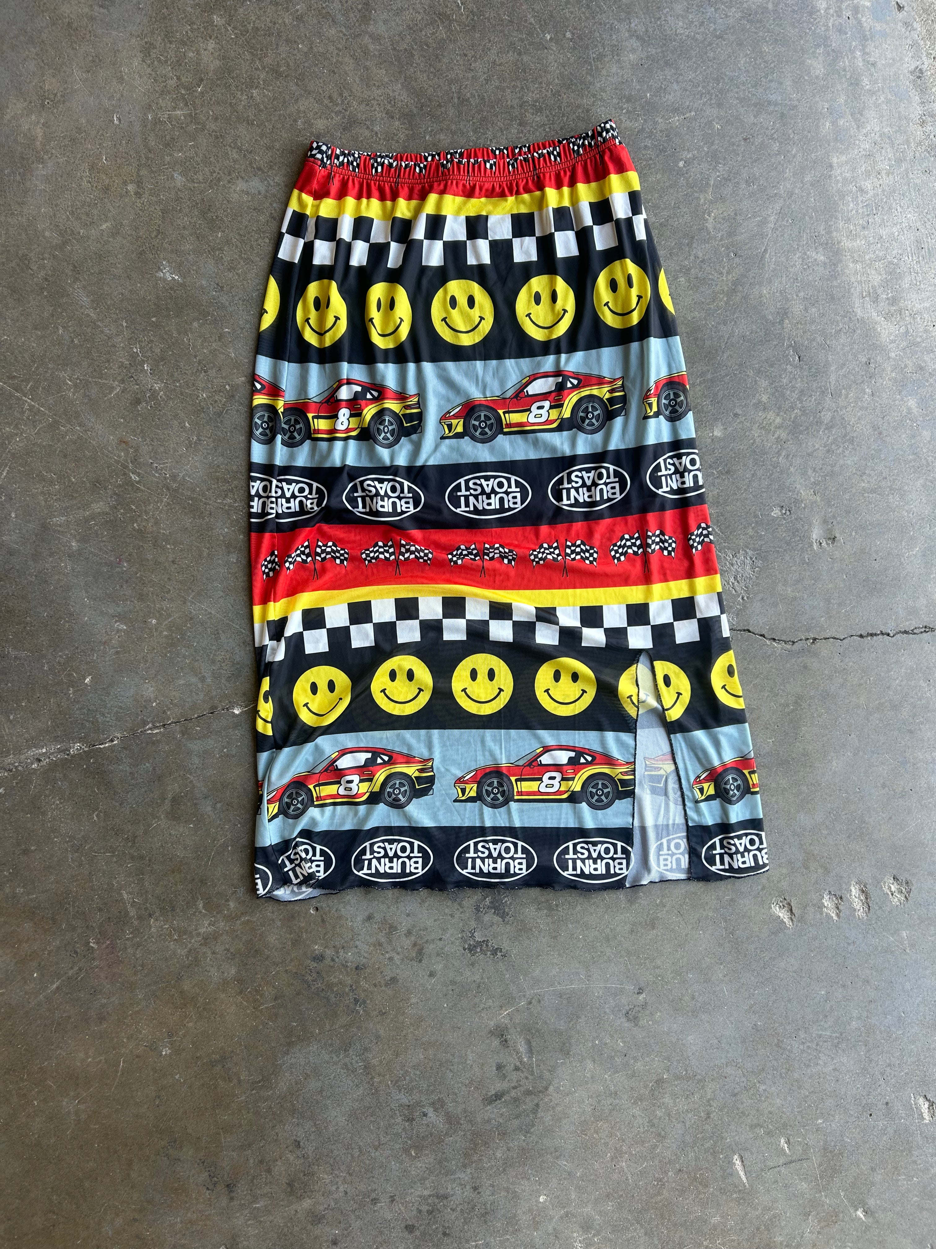 A knee-length skirt with an elastic waistband, featuring a colorful racing-themed print with yellow smiley faces, red race cars, checkered flags, and 'I'll Never Love' text, laid flat on a gray concrete floor