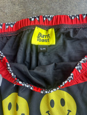 Close-up of a children's garment interior showing a yellow Burnt Toast brand label, black fabric lining, red elastic trim, and a smiley face print on the front