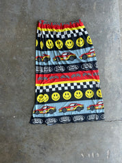 A women's maxi skirt with a colorful racing-themed print, laid flat on a gray concrete floor. The pattern features yellow smiley faces, checkered stripes, red and yellow racing cars, and 'Speed Way' logos