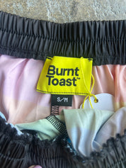 Close-up of a garment's waistband area showing a bright yellow Burnt Toast brand label, black elastic trim, and pastel pink and light green fabric layers