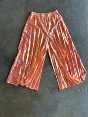 Metallic rose gold wide-leg cropped pants with a shiny, crinkled texture, laid flat on a dark gray concrete surface
