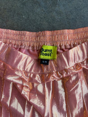 Close-up of a pink metallic ruffled garment with a visible Burnt Toast brand label at the waistband, laid flat on a dark textured surface