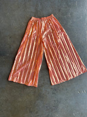 Metallic copper-orange pleated wide-leg pants laid flat on a dark gray concrete surface