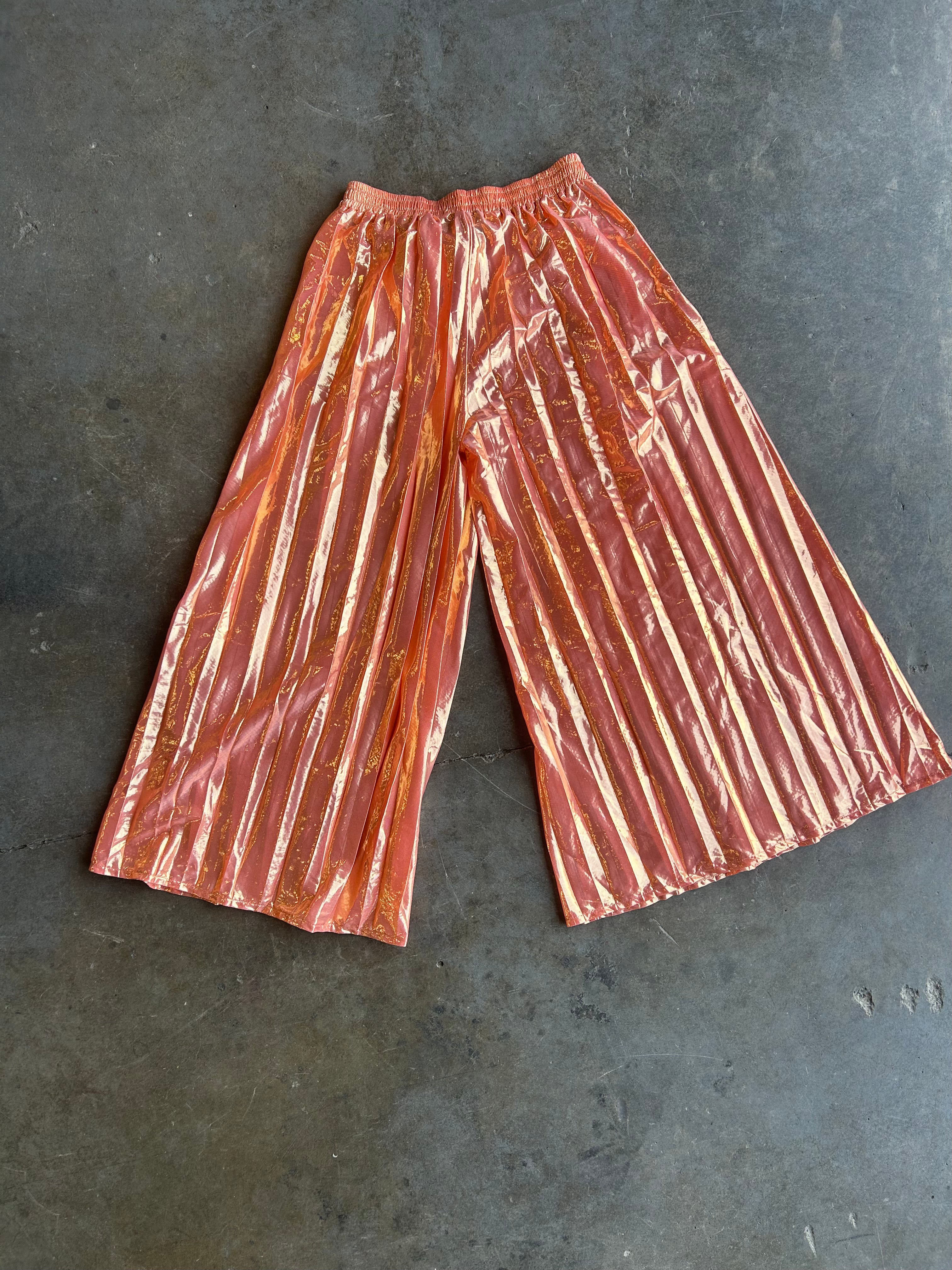 Metallic copper-orange pleated wide-leg pants laid flat on a dark gray concrete surface