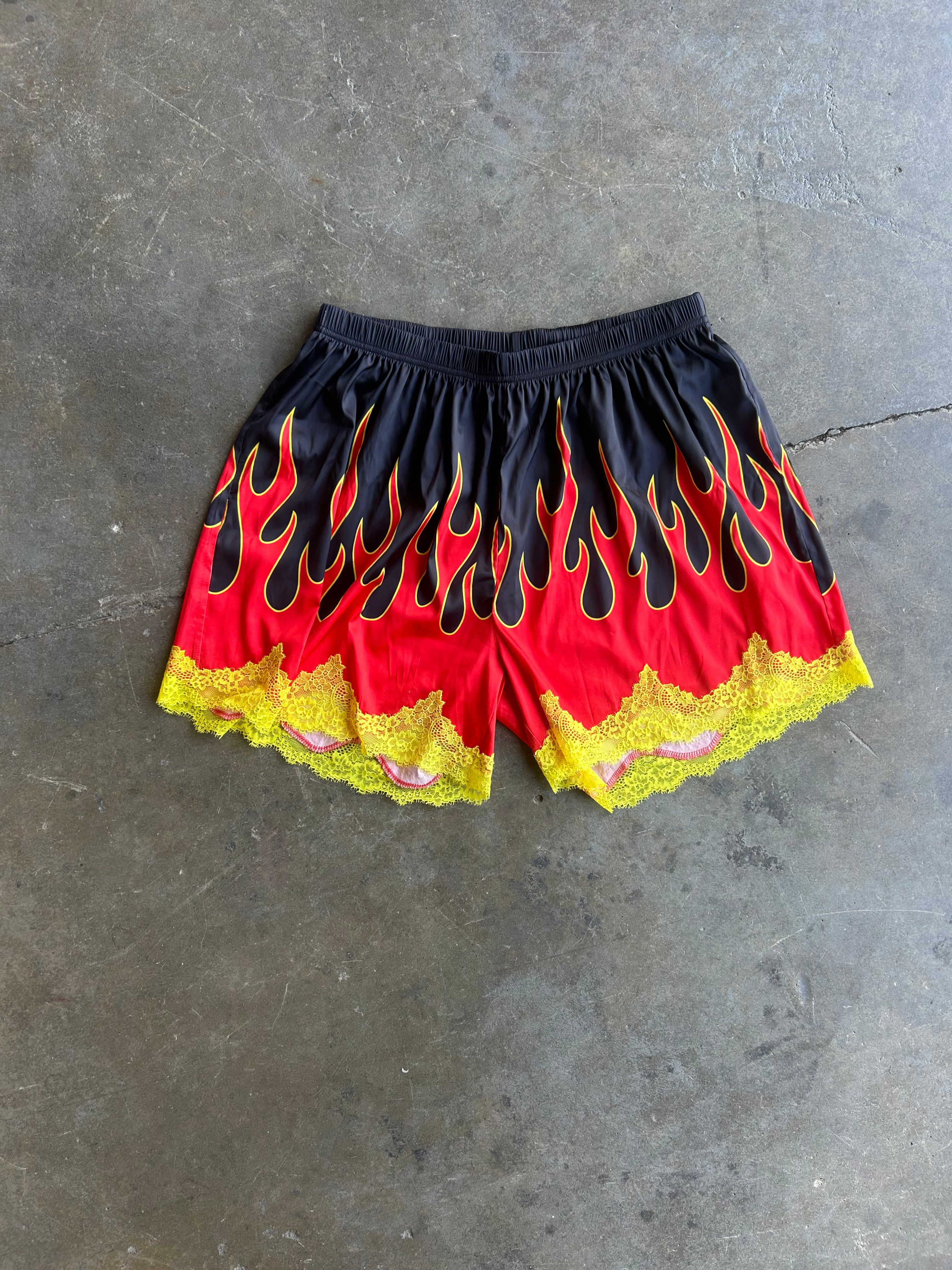 Black and red flame-print shorts with yellow lace trim, laid flat on a gray concrete floor