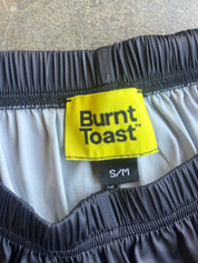 Close-up of the waistband of Burnt Toast shorts, showing a bright yellow brand label with black text, a black elastic waistband, and a light gray inner lining