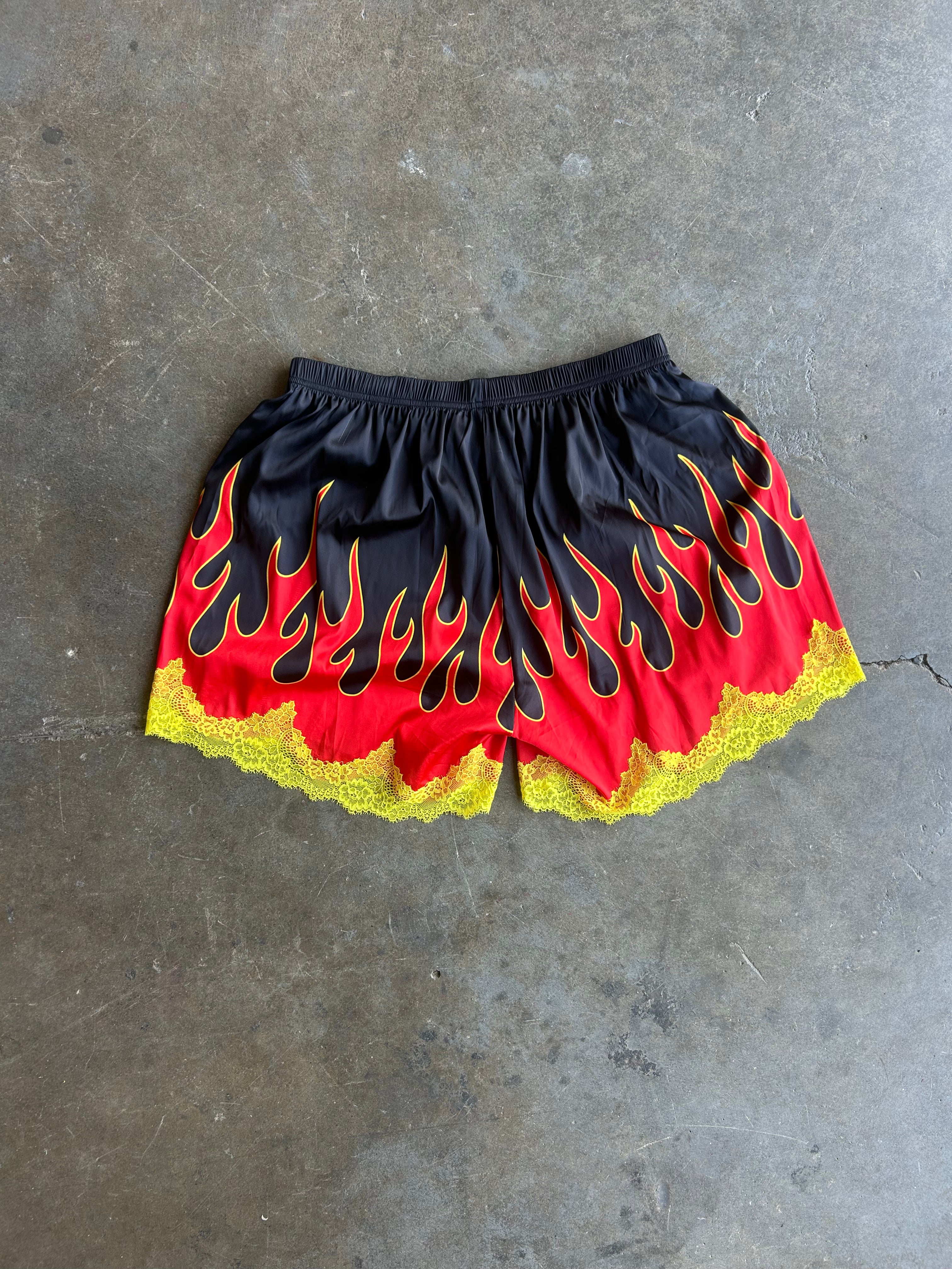 Black athletic shorts with a red and yellow flame print and yellow lace trim, laid flat on a gray concrete floor