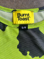 Close-up of a Burnt Toast garment neck label reading S/M, showing a neon yellow brand patch and a black-and-lime camouflage knit fabric