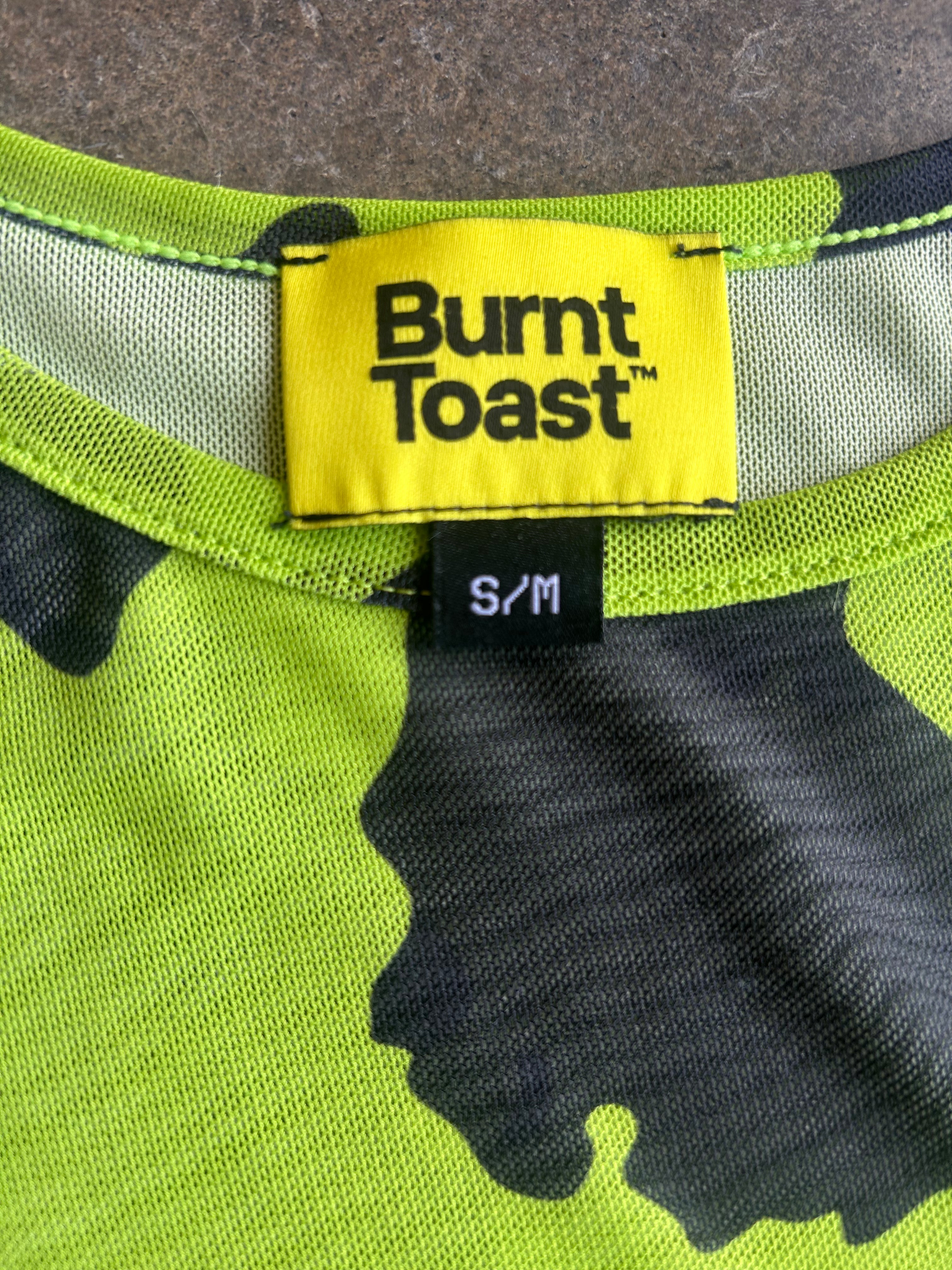 Close-up of a Burnt Toast garment neck label reading S/M, showing a neon yellow brand patch and a black-and-lime camouflage knit fabric