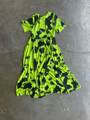 A short-sleeve midi dress with a bright neon yellow-green and black organic splatter pattern, laid flat on a gray concrete floor