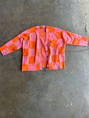 A pink and orange checkered cardigan sweater with long sleeves and front pockets, laid flat on a gray concrete floor